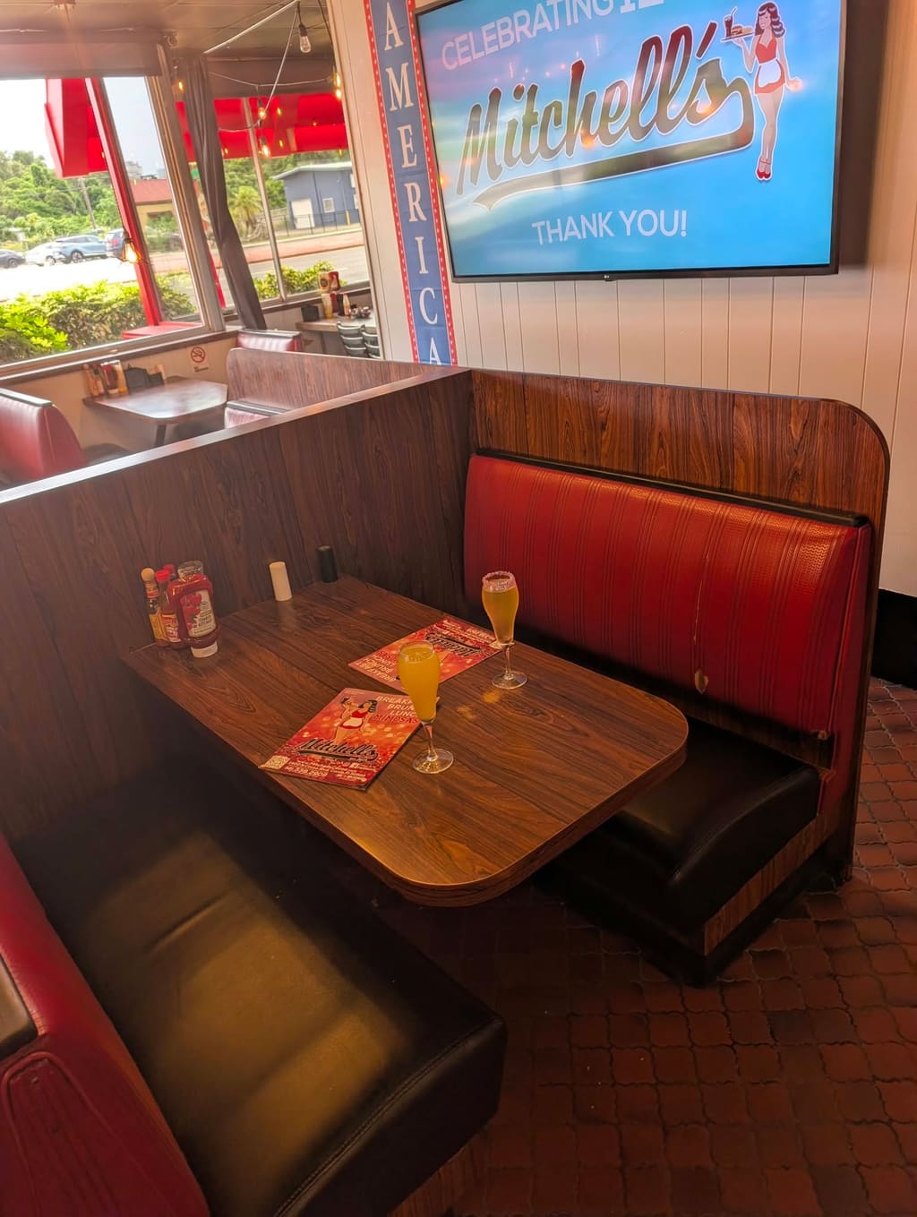 A red vinyl booth at Mitchell's with two mimosas on the table and a screen celebrating the diner's years with a thank you